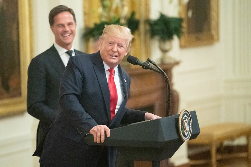 Mark Rutte laughing in the background as Donald Trump leans over to the side of the White House podium.