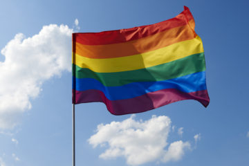 A rainbow flag flying against a cloudy blue sky