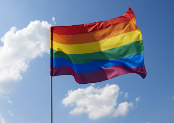 A rainbow flag waving in a cloudy blue sky