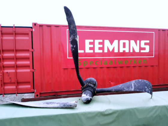 The propellor of the plane, with one blade broken off, laid out on a table in front of a red Leemans Speciaalwerken cargo container.