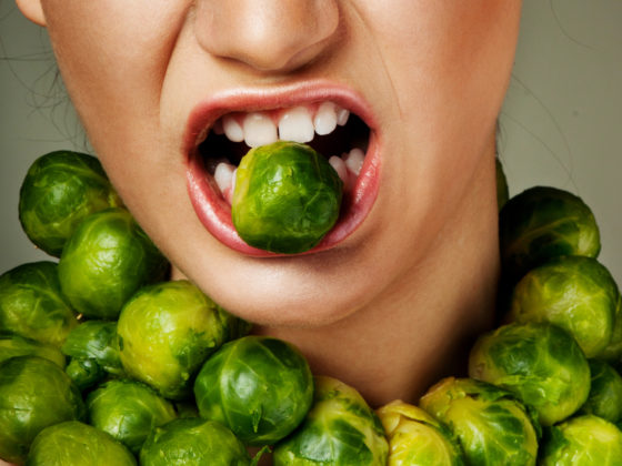 A Brussels sprout in the mouth of a woman who has more rings of sprouts around her neck